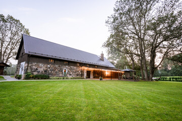 Blome, Latvia - September 11, 2023 - A scenic view of a rustic stone building with a modern roof, surrounded by lush green grass and trees, with outdoor seating and string lights.