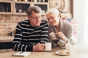 Sincere love. Happy senior couple sitting at table, drinking tea in kitchen, empty space, sun flare