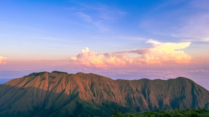 A breathtaking view of the mountains in Lombok at sunset, with golden light casting dramatic shadows on the rugged landscape and a sky painted with hues of blue and pink, creating a serene atmosphere.
