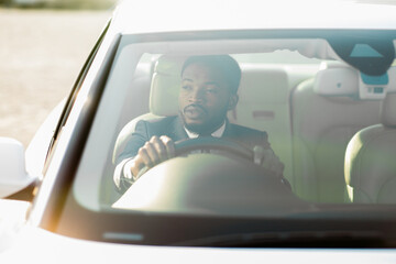 Naklejka premium Black man wearing a suit sits in the drivers seat of a car. The car is facing towards the sun, creating a bright, hazy effect. He is gripping the steering wheel and looking straight ahead