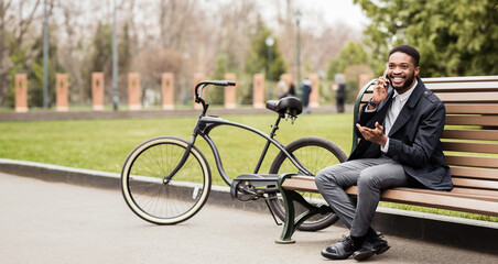 An African American man is talking on his phone in a park, sitting on a bench with his bike nearby. The setting is peaceful and green, reflecting a moment of productivity in a natural environment.
