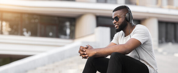 A young man wearing headphones sits on a step in an urban environment. He is wearing sunglasses and...