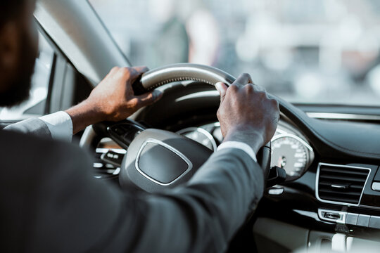A close-up image of a mans hands on the steering wheel of a car. He is wearing a suit and driving in the daytime. The background is out of focus. The driver is focused on the road.