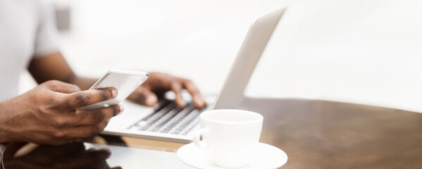 A close-up photo of a persons hands using a phone and a laptop. A coffee cup sits on a table to the...