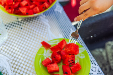 Close-up view of child's hand with fork picking up sliced piece of ripe red watermelon from plate...
