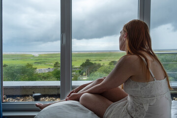 red-haired woman sits on bed gazing out large window. lush green landscape under rainy cloudy sky,...