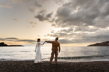 A couple standing on a beach holding hands. The sky is cloudy and the sun is setting. Scene is romantic and peaceful