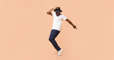 A young black man dances with a smile on his face. He is wearing a white t-shirt, blue jeans, and a black hat. He is standing against a peach background