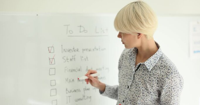 Young blonde entrepreneur ticks done tasks in to do list on white board in office slow motion. Woman builds strategy of startup project boosting