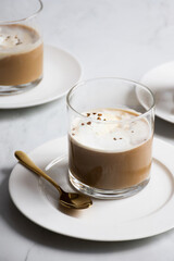 Close-up vertical shot of a glass of creamy coffee with a golden spoon on a white plate