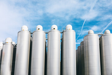 Row of tall metal tanks with a clear blue sky in the background. The tanks are silver and appear to be part of a large industrial facility