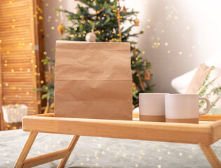 . Paper bag, kraft delivery package, tea cups, mugs on wooden tray on bed, Christmas holiday vibe.
