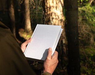 . Male hands holding book cover, blank hardcover mockup in nature.