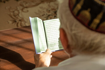 Obraz premium Lifestyle portrait of senior islamic man reading Quran at home. Natural aesthetic light