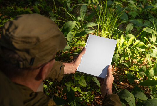. Man holding book hard cover mockup, blank hardcover mock up in nature, green plants, grass, sunlight.