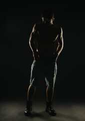 Moody portrait of a shirtless man in denim shorts with dramatic lighting capturing a silhouette in a dark room.