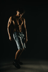 A muscular shirtless man wearing denim shorts and boots poses confidently in a dark studio setting with dramatic lighting.
