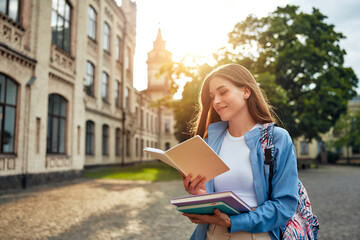 A young woman reads a book outdoors near a university at sunset, surrounded by serene nature