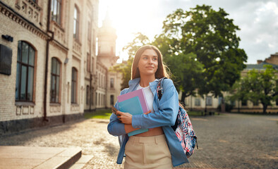 A young woman student walking on campus with a backpack and books, enjoying the sunshine