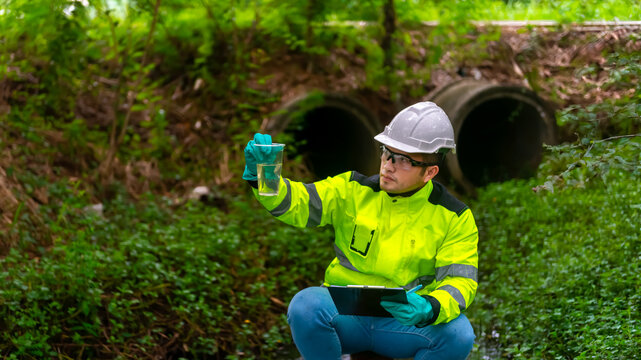 An ecologist in a green jacket sit aside of the pond looking at a liquid in a plastic glass in his hand try to examination the quality of the water. and write down the result on a clipboard