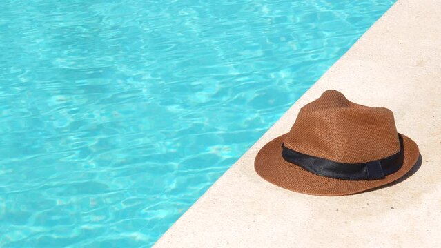 A hat is sitting on the edge of a pool. The hat is brown and black. The water is blue