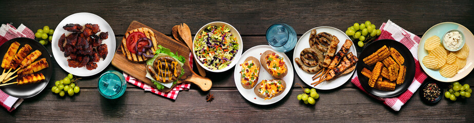 Summer BBQ or picnic food table scene. Burgers, grilled meats, potatoes, corn ribs, fruit, salad and snacks. Top view on a dark wood banner background.