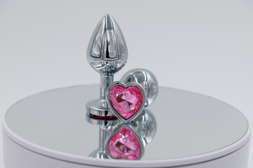 Close-up side view of two metallic butt plugs with pink heart shaped crystal lying on mirror...