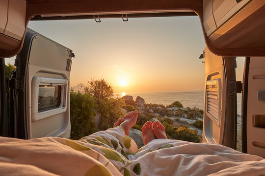 Couple enjoying a sunset view from their camper van bed, with toes emerging from under the covers, overlooking the sea. Perfect for travel, adventure, and romantic getaway themes.
