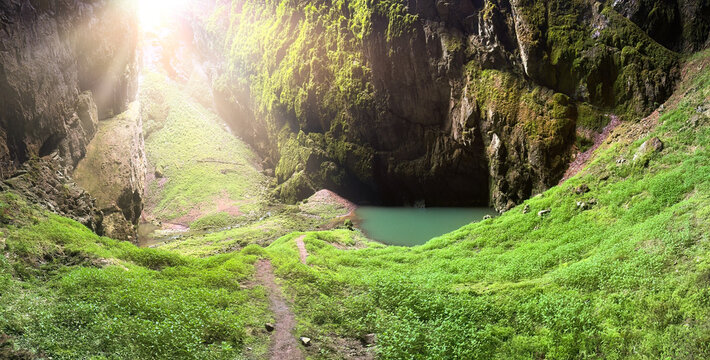 Panoramic view of Macocha Abyss in Moravian Karst, Czech Republic. A lush, green valley inside a deep sinkhole, illuminated by sunlight streaming through the opening. Vibrant vegetation and pond.