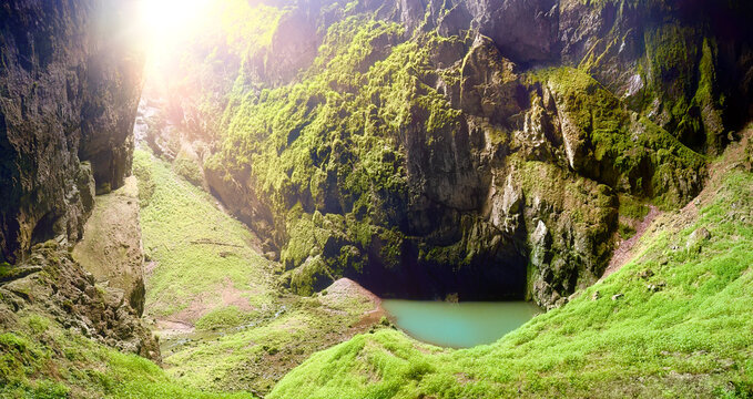Panoramic view of Macocha Abyss in Moravian Karst, Czech Republic. A lush, green valley inside a deep sinkhole, illuminated by sunlight streaming through the opening. Vibrant vegetation and pond.