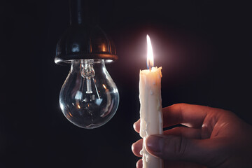 Burning candle near a switched off light bulb in complete darkness. Blackout, electricity off, energy crisis or power outage, concept image.