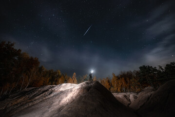 A man stands on a hill and points a flashlight at the starry night sky. Mountain climbing. Standing on a rocky peak Silhouette of a climber or tourist.