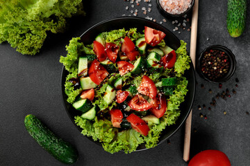 Vegetable salad of tomatoes, cucumbers, onions, lettuce dressed with aromatic dressing in a black plate on a gray background