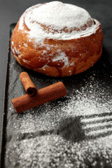 Round, delicious bun with cinnamon and powdered sugar on a black plate on a gray background