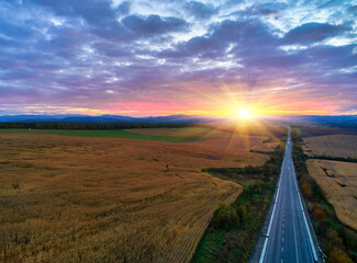 Aerial view of mountain road at sunset in autumn. Top view from drone of road in hills. Beautiful landscape with roadway, trees, green meadows, sky with golden sunlight in fall.