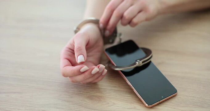 Woman tries to take off handcuffs connected with modern smartphone sitting at wooden table. Female person feels dependent on electronic device slow motion