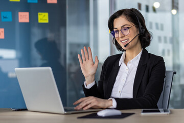 Businesswoman wearing a headset and waving during a video conference call while sitting in a modern...