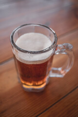 Glass mug with fresh cold beer on wooden table on patio. A refreshing summer drink. Liquor. A pint of beer. Foam beer in a glass mug. Street cafe. Summer vacation. Abnormal heat.