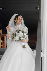A woman in a wedding dress holding a bouquet of flowers. The dress is white and has a long train. The woman is standing in front of a door, possibly getting ready to leave for her wedding