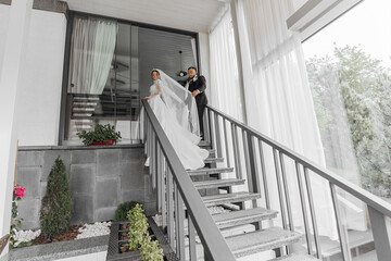 A bride and groom are walking up the stairs to their wedding ceremony. The bride is wearing a white dress and the groom is wearing a black suit. The scene is set in a home, with a potted plant