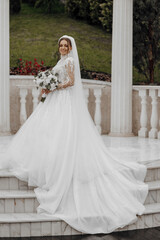 Fototapeta premium A woman in a white wedding dress is standing on a marble staircase. She is holding a bouquet of flowers in her hand. Concept of elegance and celebration, as the bride is dressed in a beautiful gown