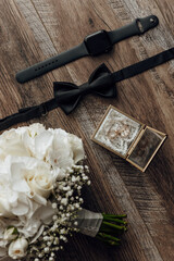 A bouquet of white flowers sits on a wooden table next to a watch and a tie. The flowers are arranged in a vase and the tie is folded