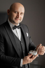 A man in a suit holding a box. The man is smiling and wearing a bow tie. The box is silver and has a shiny appearance