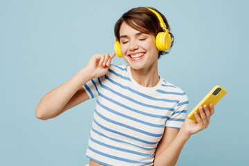 Young smiling happy fun woman wear striped t-shirt casual clothes listen to music in headphones hold use mobile cell phone isolated on plain pastel light blue cyan background studio Lifestyle concept