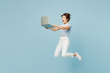 Full body side view young surprised shocked IT woman wears striped t-shirt casual clothes jump high hold use work on laptop pc computer isolated on plain light blue cyan background. Lifestyle concept.