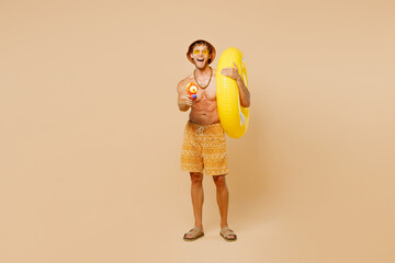 Full body young happy man wear yellow shorts swimsuit hat relax rest near hotel pool hold rubber ring play shoot from water gun isolated on plain beige background. Summer vacation sea sun tan concept.