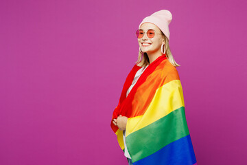 Side profile view young happy fun lesbian woman wrapped in striped rainbow flag looking aside on area isolated on plain purple wall background studio portrait. Pride day June month love LGBTQ concept.