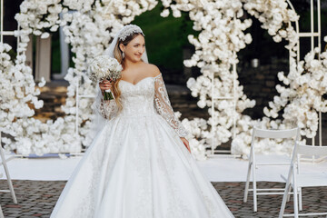 Fototapeta premium A woman in a white wedding dress is standing in front of a white archway. She is holding a bouquet of flowers and she is posing for a picture. Concept of elegance and celebration