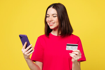 Young woman wear pink t-shirt casual clothes using mobile cell phone hold credit bank card do online shopping order delivery booking tour isolated on plain yellow orange background. Lifestyle concept.