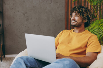 Young minded pensive Indian IT man wear orange casual clothes work hold use laptop pc computer sits in armchair stay at home hotel flat rest relax spend free spare time in living room. Lounge concept.
